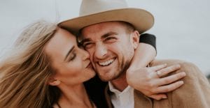 Blonde woman kisses a smiling man wearing a tan cowboy hat on