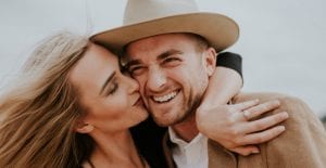Blonde woman kisses a smiling man wearing a tan cowboy hat on