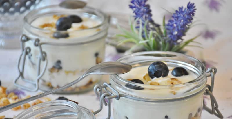 Two glass containers are full of white yogurt, granola, and blueberries next to a blue flower