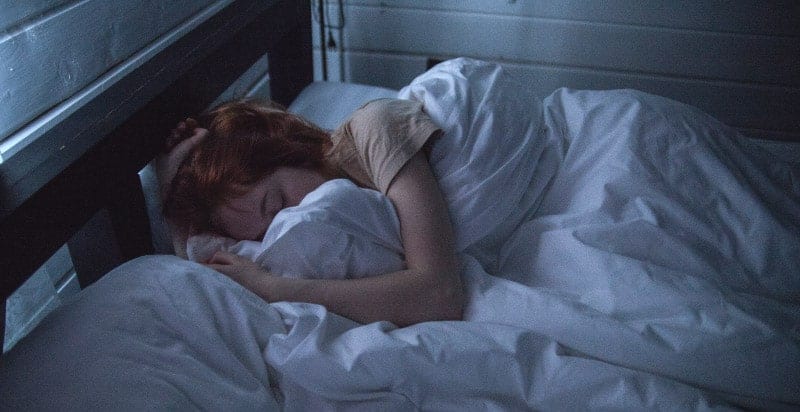 Red haired woman sleeps on her side wrapped in white sheets in a bed with a black frame