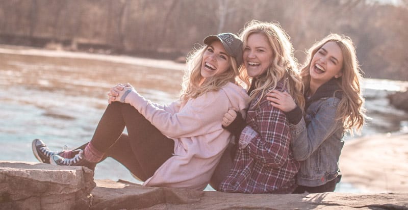 3 young women wearing warm clothing sit next to a river smiling and laughing on a sunny winter day
