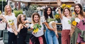 Group of young women hold flower bouquets at an outdoor market and smile with very white teeth