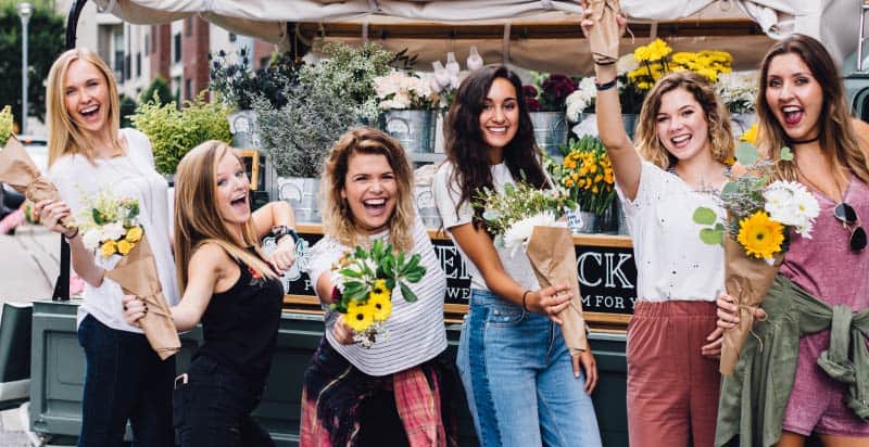 Group of 6 young women hold flower bouquets at an outdoor market and smile with lipstick and very white teeth