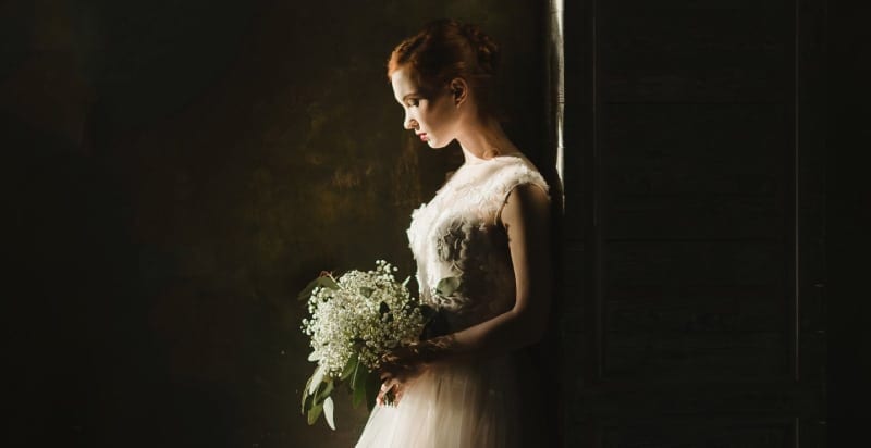 Red-haired bride in a lace dress holds a bouquet of white flowers, looking down sadly as she stands alone in the dark