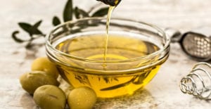 Closeup of a stream of yellow olive oil being poured into a clear bowl oil next to a cluster of green olives