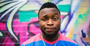 Dark-haired man wearing a pink and blue shirt in front of a wall of graffiti looks quizzical as he wonders about the dentist