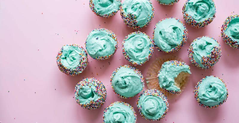 Aerial view of vanilla cupcakes with blue frosting and colorful sprinkles on a pink counter