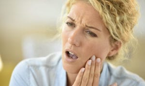 Blonde woman cringes and touches her cheek due to extreme tooth pain from dry socket