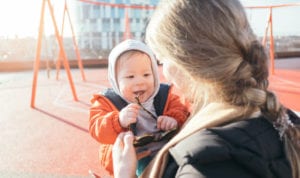 Baby wearing an orange jacket and gray beanie is held by his mother by a swingset at a playground