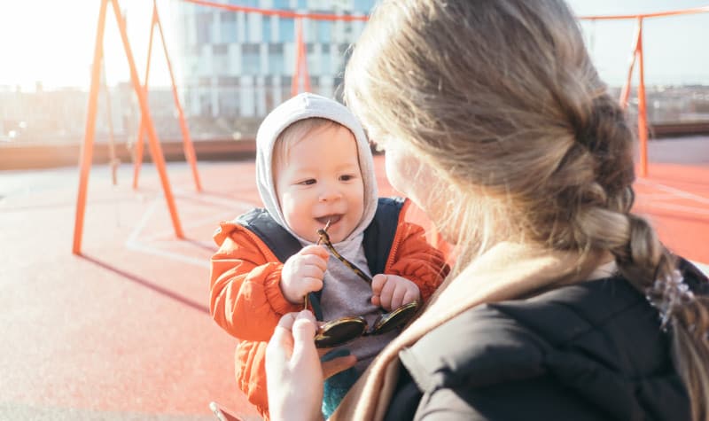 Baby wearing an orange jacket and gray beanie is held by his blonde mother by a swingset at a playground
