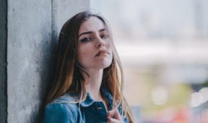 Brunette woman wearing a denim jacket stands against a stone wall looking anxious about her canker sore