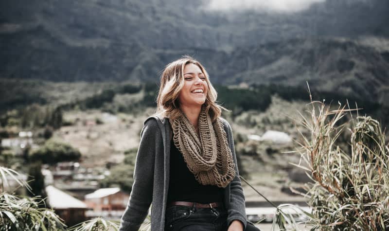 Brunette woman wearing a gray cardigan, tan scarf, black shirt, and jeans smiles while standing by a mountain